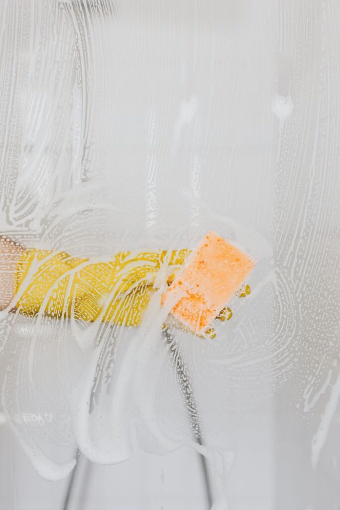 Crop anonymous person in yellow latex protective glove washing glass shower partition in modern bathroom using detergent and sponge
