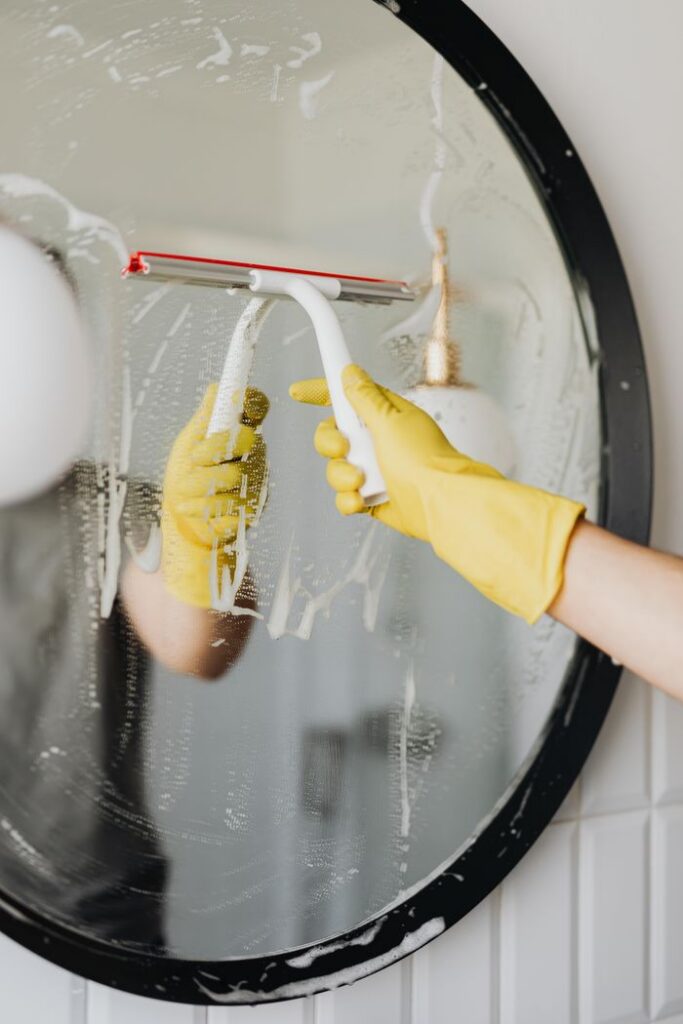 Faceless housekeeper in yellow rubber gloves cleaning round mirror from foam by squeegee in bathroom